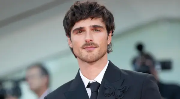 Jacob Elordi style: en ung man med mörkt hår och mustasch bär en svart kostym med en fjärilsbrosch.