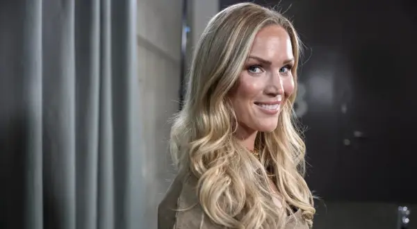 Marie Serneholt ler mot kameran med sitt blonda hår fallande i lockar. Hon bär en beige jacka och ett guldigt halsband.
