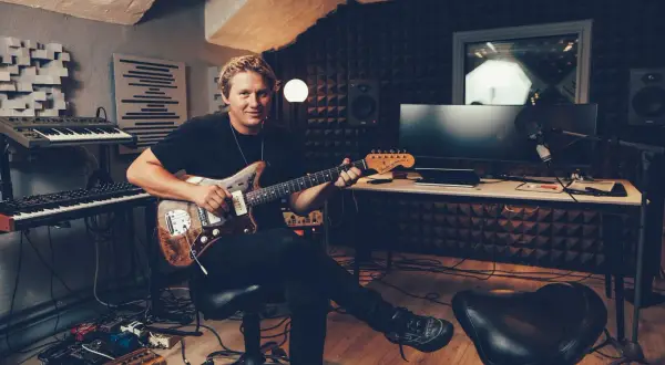 John Engelbert sitter i en inspelningsstudio med en gitarr i famnen, omgiven av musikinstrument och teknisk utrustning.