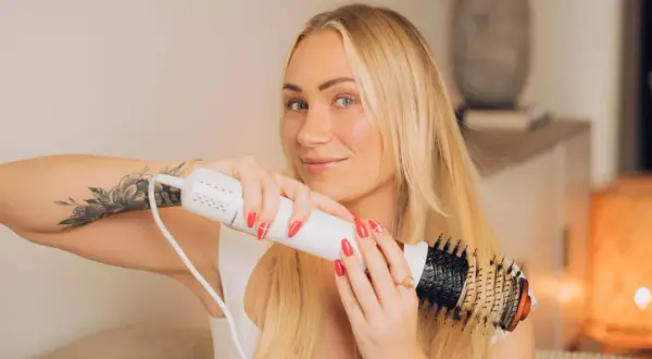 Blond kvinna med tatuering på armen stylar sitt hår med en varmluftsborste för perfekt blow out hår.