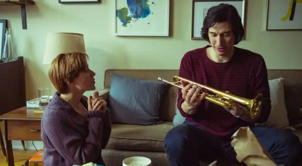 Adam Driver håller i en trumpet i en scen från en av hans filmer. Bredvid honom sitter en kvinna och tittar på.