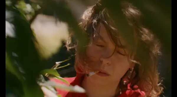 En ung kvinna med slutna ögon och en cigarett i munnen, omgiven av grönska. Scenen påminner om stämningen i sorgliga filmer.