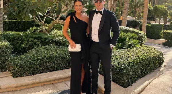 Ett par klädda för black tie dresscode, hon i en elegant svart klänning, han i smoking. De poserar glatt utomhus bland grönska.