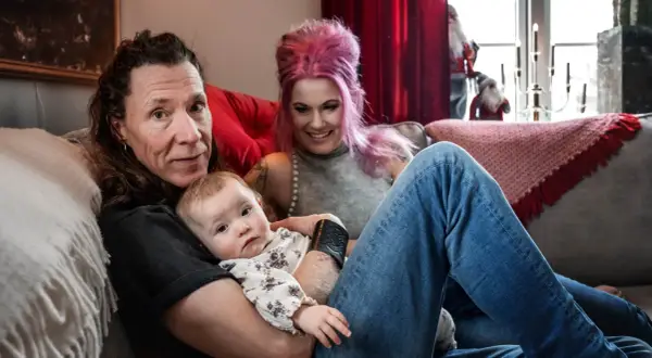 En familj med ett etype barn myser i soffan. Pappan håller barnet, mamman med rosa hår ler.