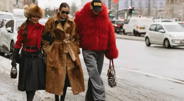 Tre kvinnor i trendiga ytterkläder, en med röd pälsjacka, visar upp **copenhagen street style** på en snöig gata.
