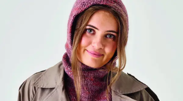 Ung kvinna med blå ögon bär en stickad balaclava i rosa och lila nyanser, med en beige trenchcoat.