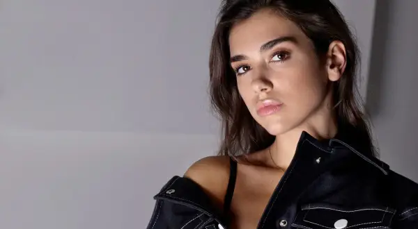 Dua Lipa utan smink, med mörkt hår och en mörk jacka som sitter löst över axeln.