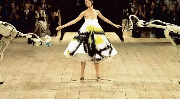 Shalom Harlow ung i en vit klänning med svart och gul spraymålning, omgiven av robotarmar på en catwalk.