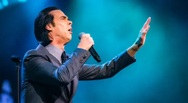 Nick Cave sjunger passionerat med mikrofonen i handen under en nick cave tour. Hans arm är utsträckt mot en blå bakgrund.
