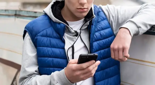Ung kille med blå dunjacka och grå hoodie kollar på sin mobil. Kanske undrar han vad betyder simp?
