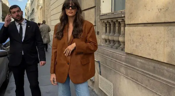 En kvinna bär en snygg, brun vero moda mockajacka och jeans på en stadsgata. En livvakt går bakom henne.