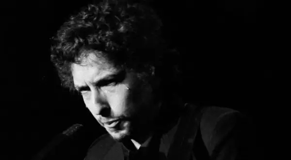 Bob Dylan, ikoniskt ansikte i en svartvit film, med lockigt hår och intensiv blick.