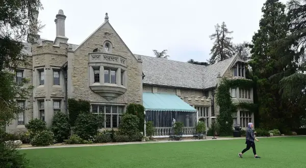 En person går över den gröna gräsmattan framför Playboy Mansion, en ikonisk byggnad med stenfasad och tak av skiffer.
