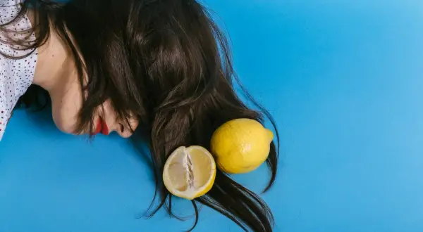 En person med mörkt hår och röda läppar ligger på en blå bakgrund med en hel och en halv citron. Kanske en idé för att bleka håret med citron?