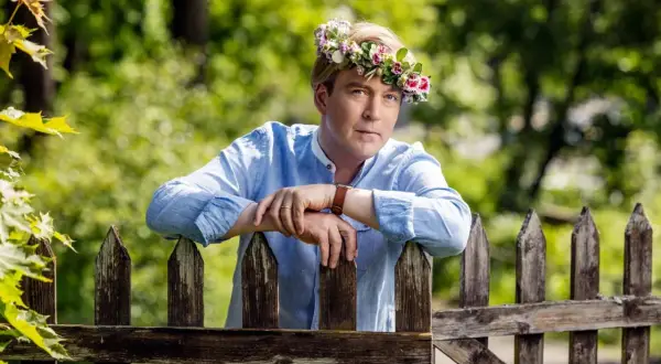 En man med blomsterkrans på huvudet lutar sig mot ett staket. En sommarprat recension som fångar essensen av svensk sommar.
