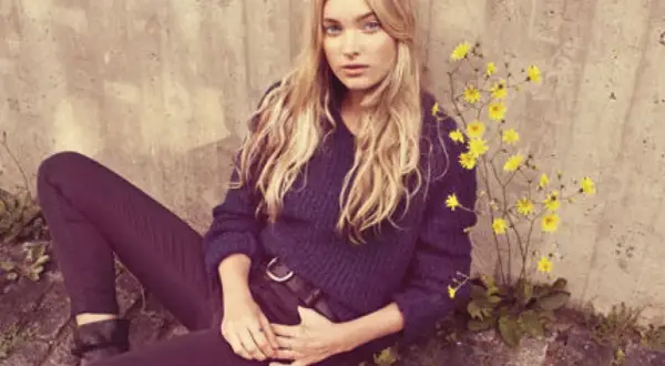 Elsa Hosk, med sitt blonda hår och blå ögon, sitter avslappnat mot en betongvägg, omgiven av gula blommor.