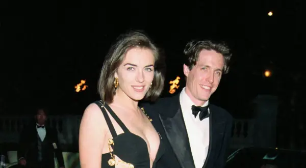 Elizabeth Hurley 90s i Hugh Grant ler mot kameran. Hon bär en ikonisk svart klänning med gulddetaljer.