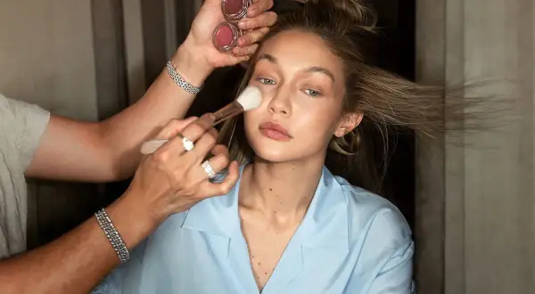 En makeupartist applicerar puder med en borste på en modell. Två små burkar med rouge syns i handen. En perfekt finish för baking makeup.