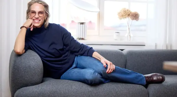 Tyra Wiklund, med ett leende, vilar på en grå soffa i jeans och mörkblå tröja.