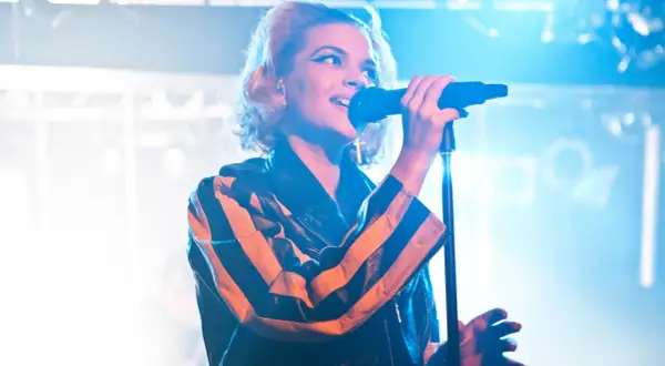 En artist med tove styrke sjunger i mikrofonen, iklädd en läderjacka med orange och svarta ränder.