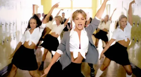 En grupp dansare, med fokus på en blond kvinna i mitten, utför en koreografi i en skolkorridor. Deras **blonde halloween costumes** är inspirerade av skoluniformer.