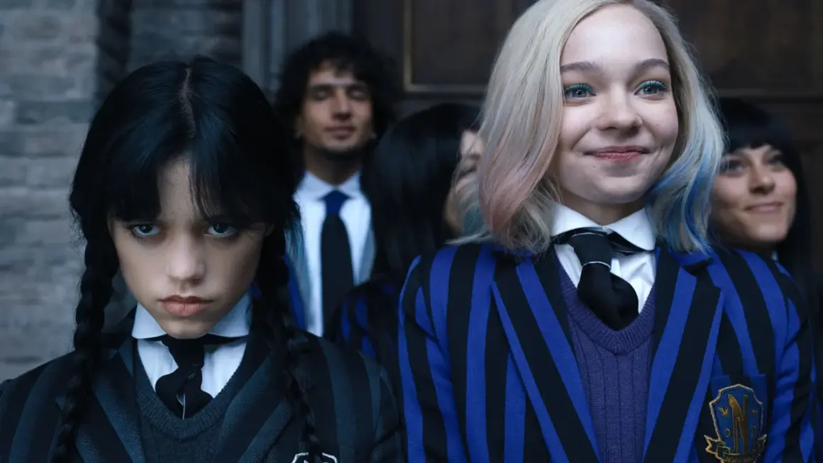Skådespelare Wednesday Addams med flätor och en allvarlig min, bredvid en leende elev med blont hår och blåa inslag.
