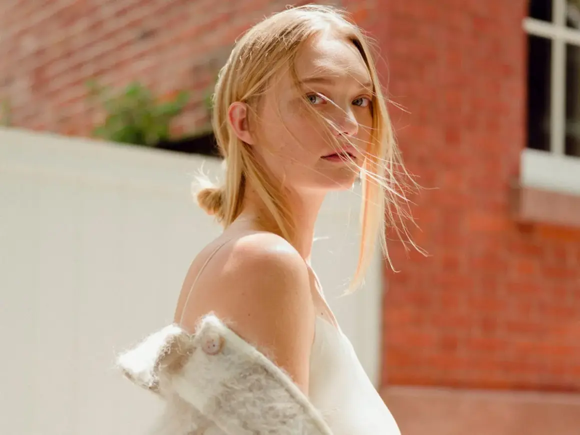 Gemma Ward med blont hår och en vit klänning, med en ullig jacka över axlarna, står framför en tegelvägg.