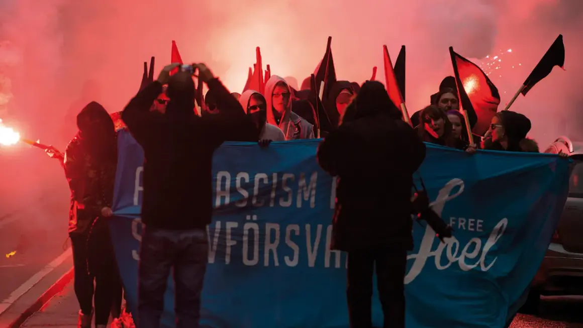 En grupp demonstranter med facklor och banderollen "ASCISM FÖRSVANN" och "FREE Joel". De agerar som en anti-hjälte, som kämpar mot orättvisor.