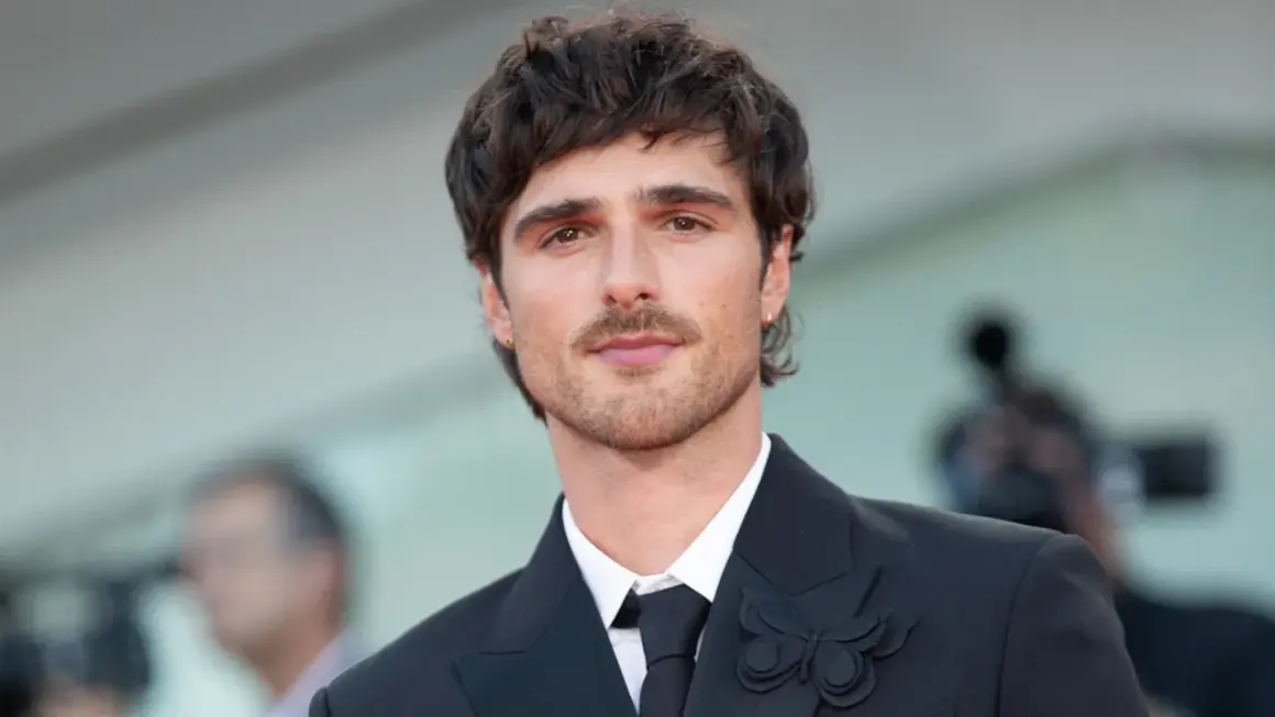 Jacob Elordi style: en ung man med mörkt hår och mustasch bär en svart kostym med en fjärilsbrosch.