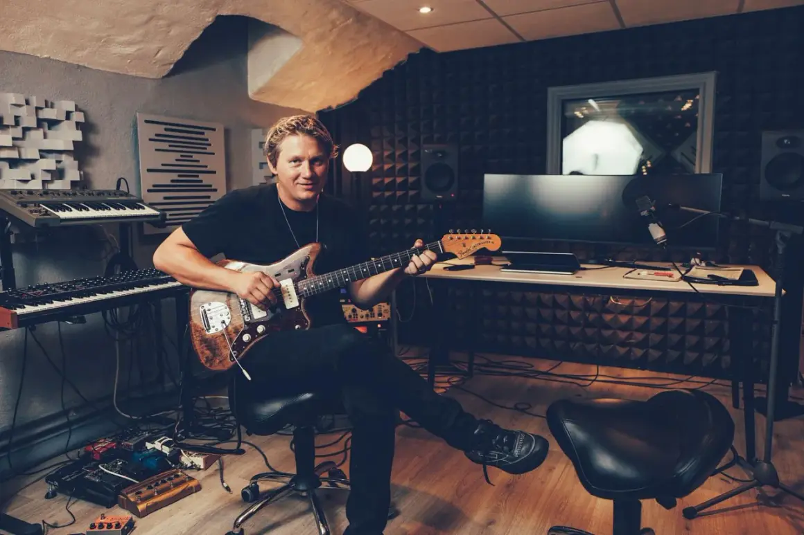 John Engelbert sitter i en inspelningsstudio med en gitarr i famnen, omgiven av musikinstrument och teknisk utrustning.