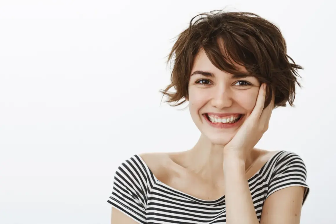 Glad kvinna med en lekfull jellyfish haircut ler brett, handen mot kinden.