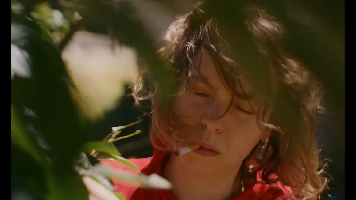 En ung kvinna med slutna ögon och en cigarett i munnen, omgiven av grönska. Scenen påminner om stämningen i sorgliga filmer.