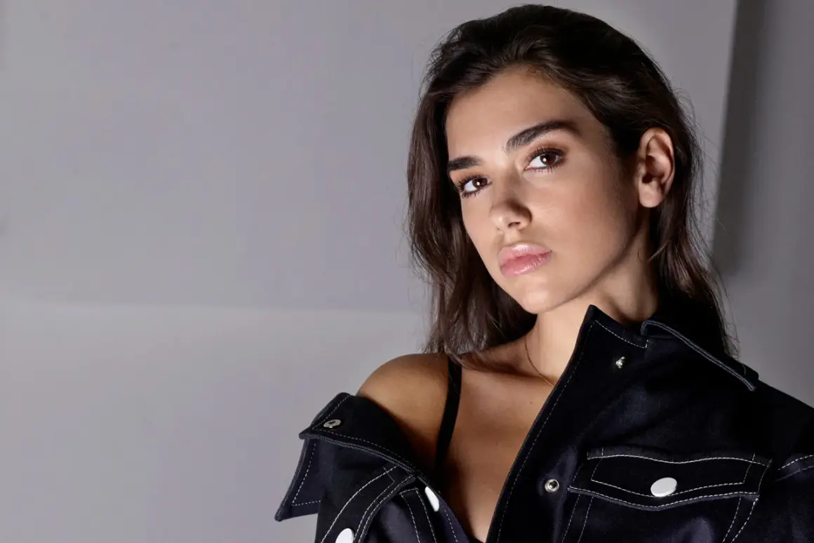 Dua Lipa utan smink, med mörkt hår och en mörk jacka som sitter löst över axeln.