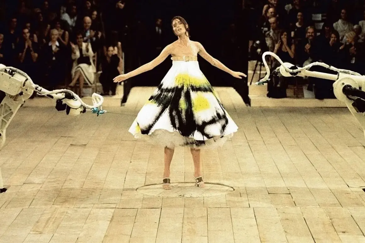 Shalom Harlow ung i en vit klänning med svart och gul spraymålning, omgiven av robotarmar på en catwalk.