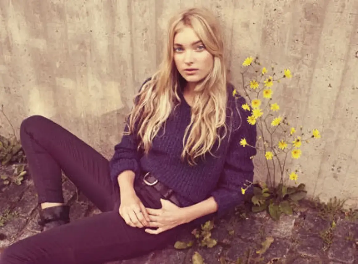Elsa Hosk, med sitt blonda hår och blå ögon, sitter avslappnat mot en betongvägg, omgiven av gula blommor.