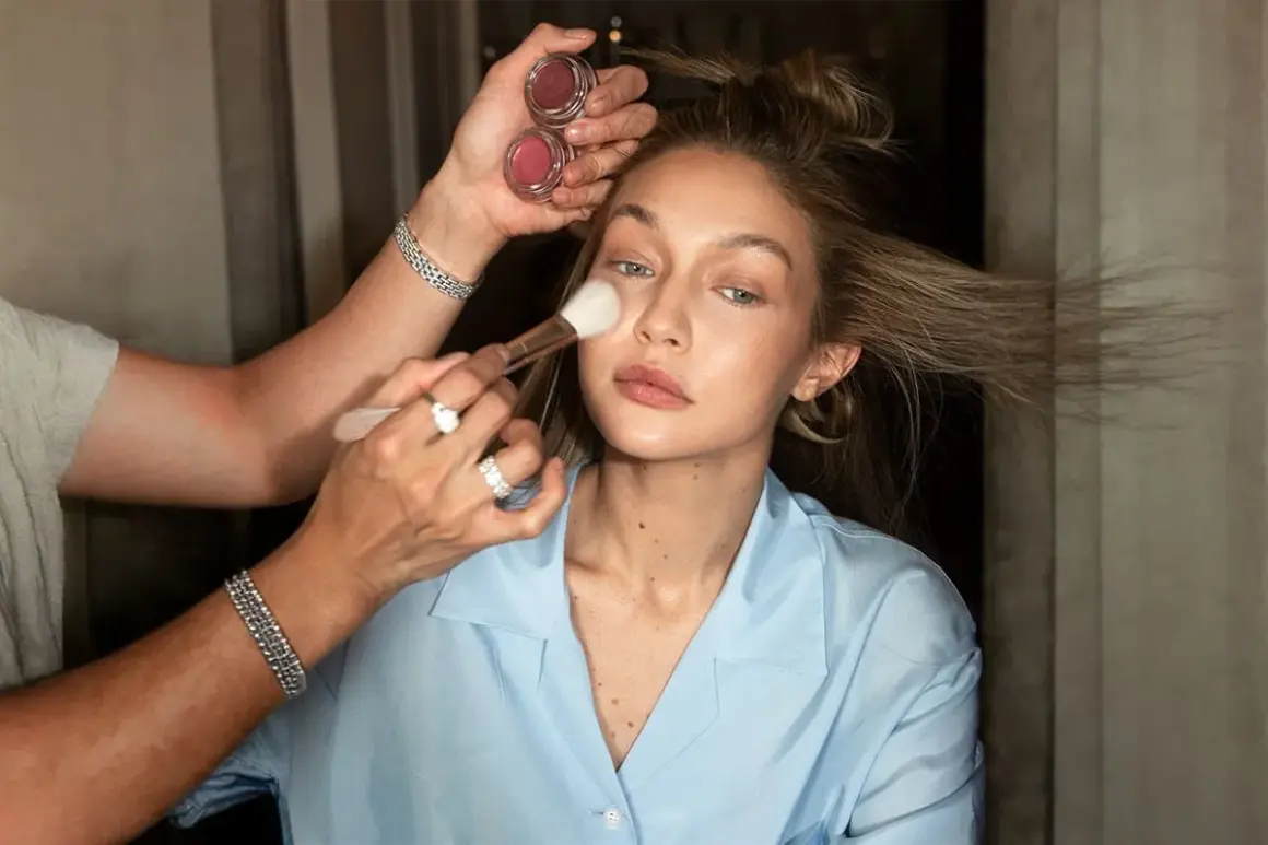 En makeupartist applicerar puder med en borste på en modell. Två små burkar med rouge syns i handen. En perfekt finish för baking makeup.