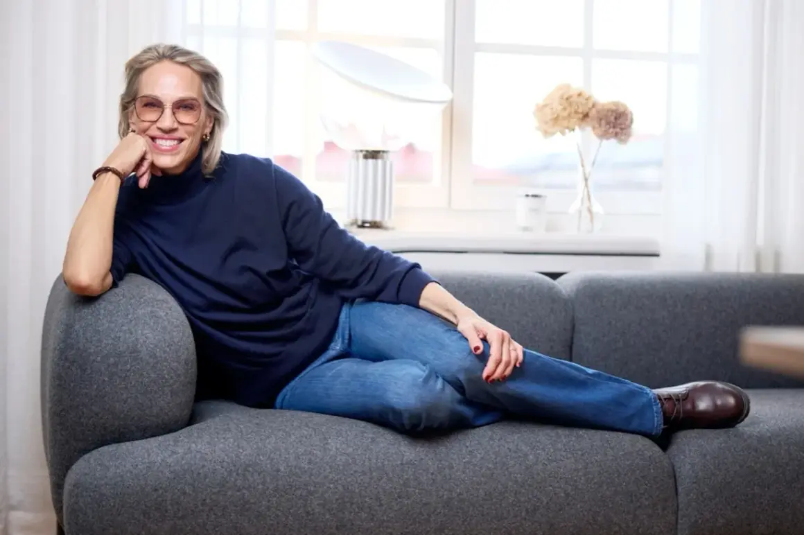 Tyra Wiklund, med ett leende, vilar på en grå soffa i jeans och mörkblå tröja.