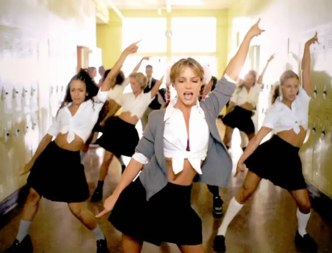 En grupp dansare, med fokus på en blond kvinna i mitten, utför en koreografi i en skolkorridor. Deras **blonde halloween costumes** är inspirerade av skoluniformer.