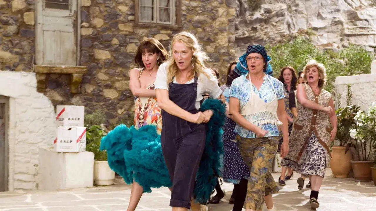 Kvinnor i glada kläder springer, som i Mamma Mia 3. En med turkos volanger, en i blå overall.