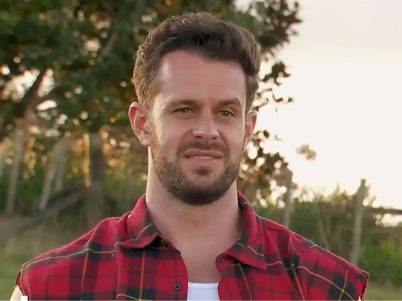 Bachelor Simon, med rufsigt hår och rutig skjorta, ler mot kameran utomhus.