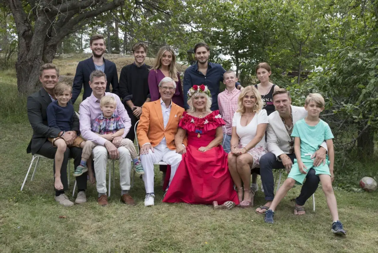 Stor familj samlad utomhus. I mitten sitter en kvinna med blomsterkrans, omgiven av sina barn och barnbarn. En av dem är Hans Wahlgrens barnbarn.
