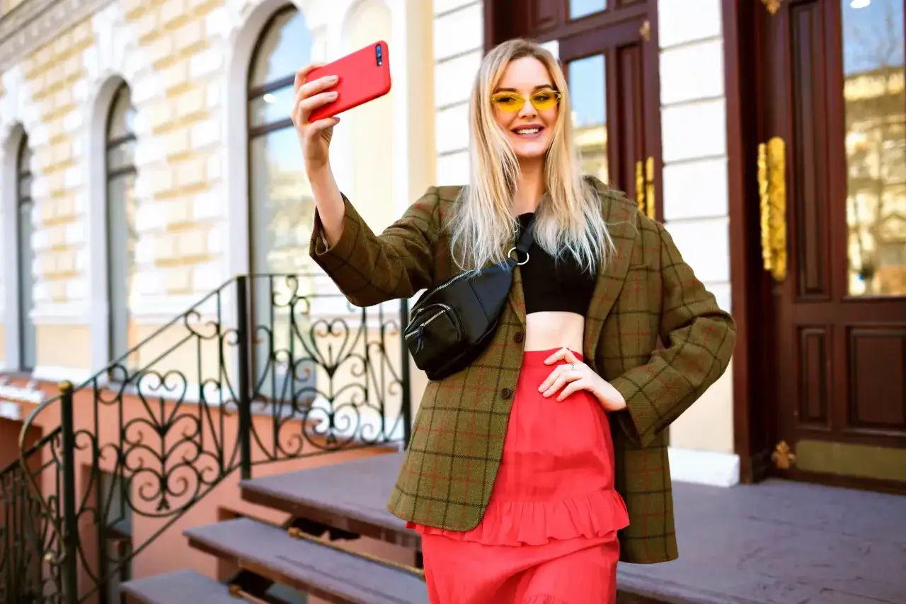 En kvinna med blont hår och gula solglasögon tar en selfie med en röd telefon. Hon bär en rutig kavaj, en svart topp och en röd kjol, en outfit som speglar hennes mimetic desire för trendiga kläder.