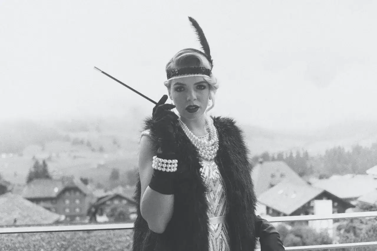 En kvinna i 1920-talsmode med fjäderprydd pannband, pärlhalsband och en cigarettmunstycke.