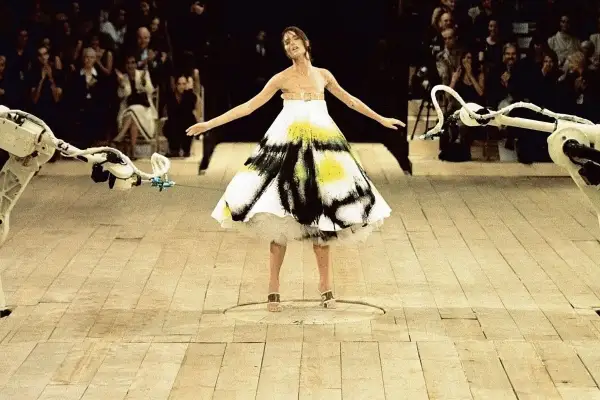 Shalom Harlow ung i en vit klänning med svart och gul spraymålning, omgiven av robotarmar på en catwalk.