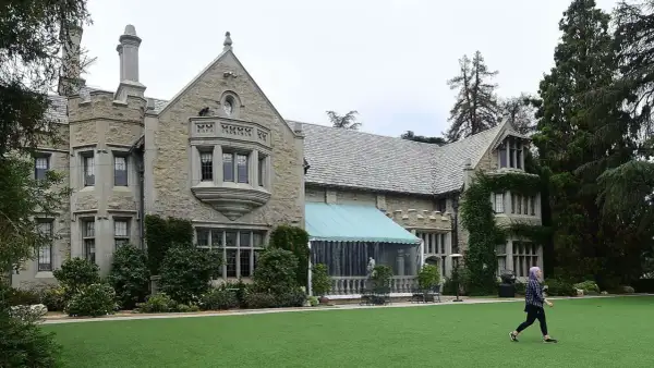 En person går över den gröna gräsmattan framför Playboy Mansion, en ikonisk byggnad med stenfasad och tak av skiffer.