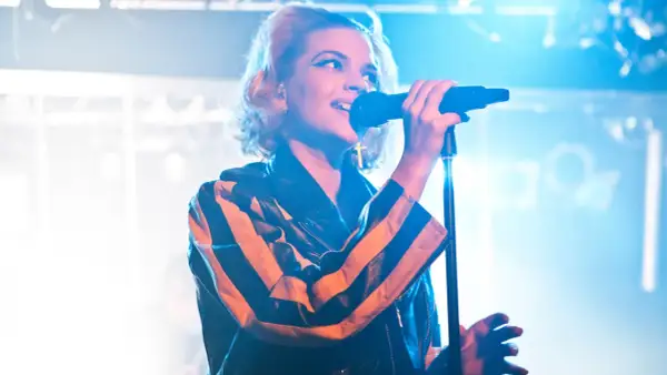 En artist med tove styrke sjunger i mikrofonen, iklädd en läderjacka med orange och svarta ränder.